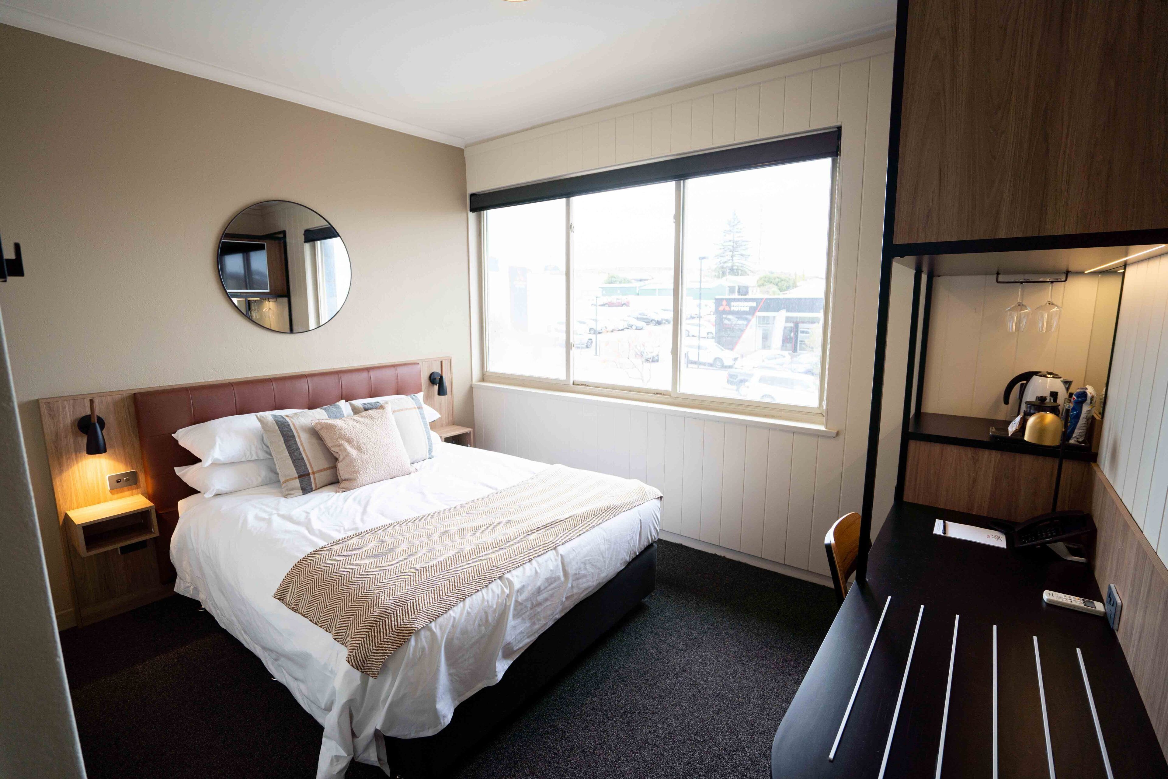 A hotel room with a large bed, a bedside table with a lamp, a round mirror, a window with a view of the outside, and a desk with a chair.