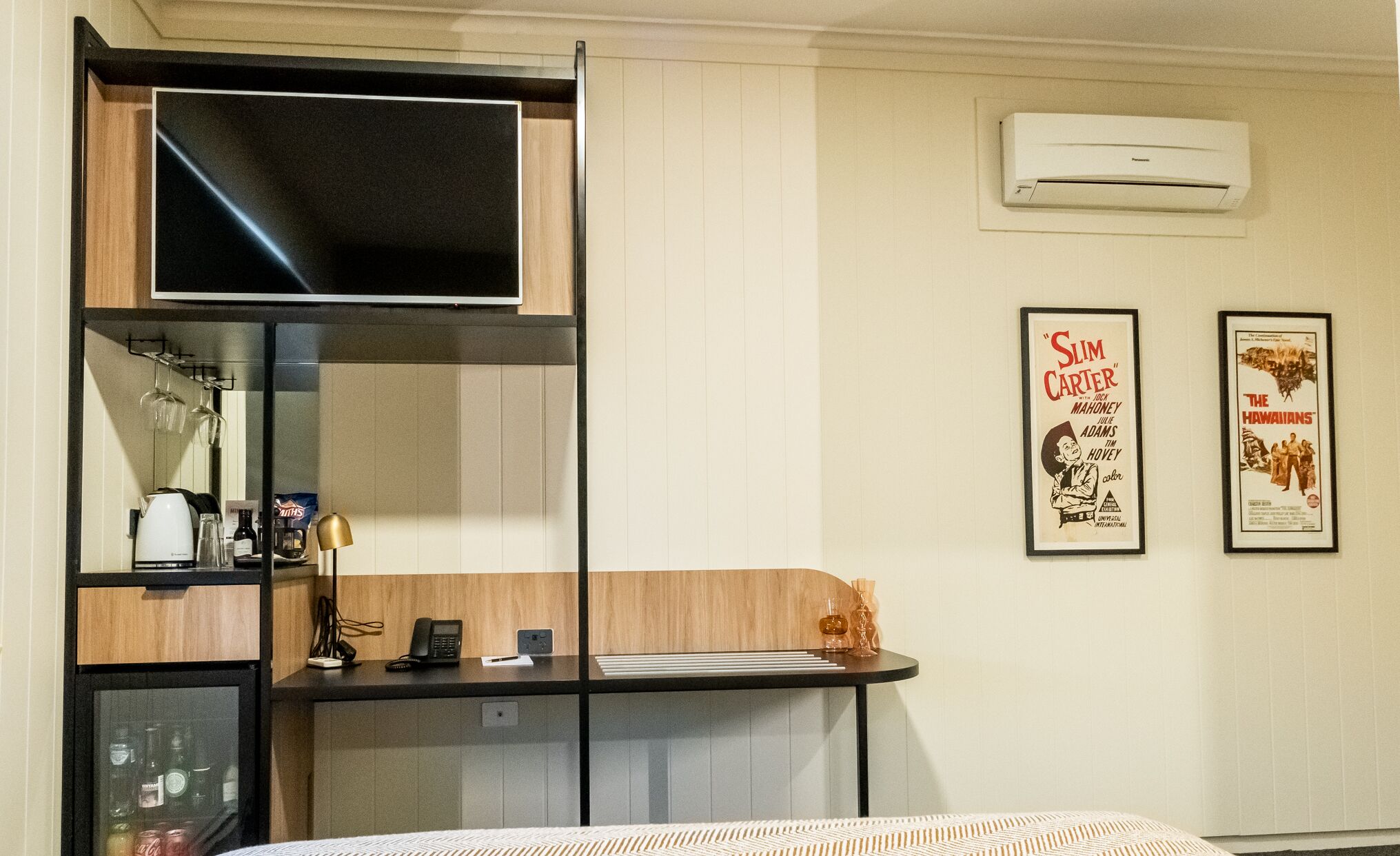 A hotel room with a bed, a desk with a computer, a minibar, a television, and two framed movie posters on the wall.
