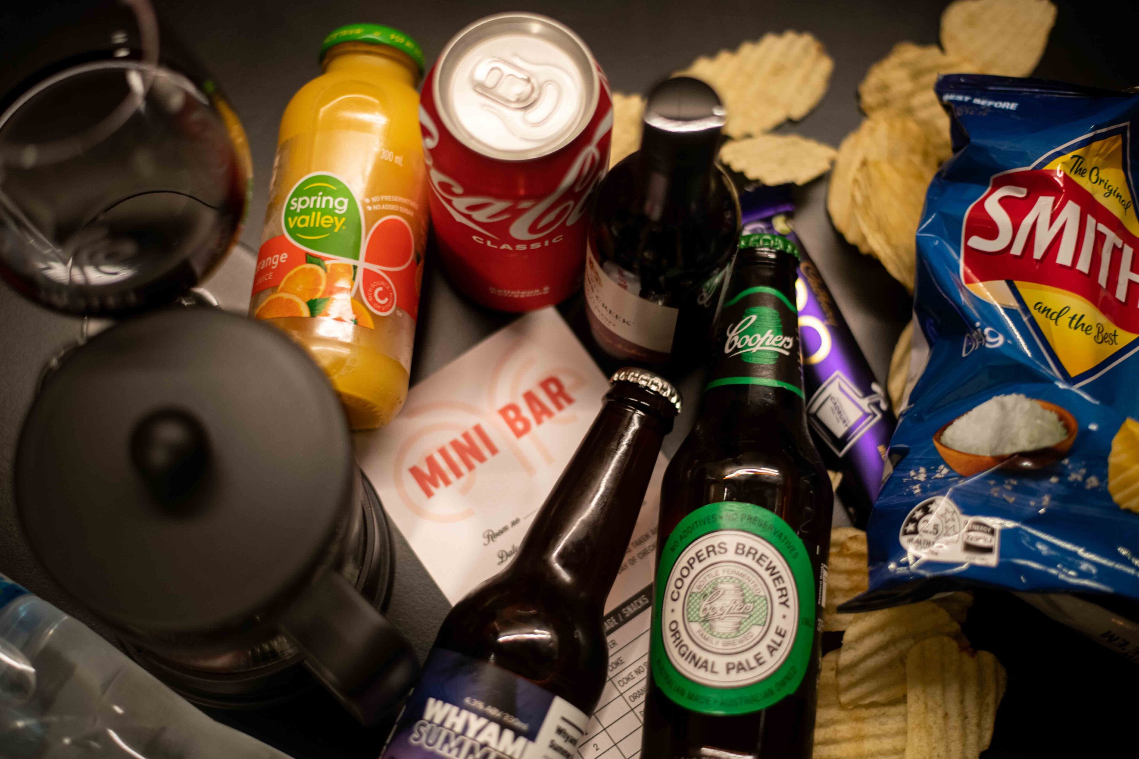 Una varietà di cibi e bevande, tra cui una lattina di Coca-Cola, una bottiglia di Coors Brewery Original Pale Ale, una bottiglia di Whyam Summer Ale, una busta di patatine Smith e un menu del minibar.