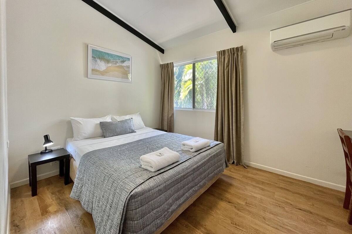 Ein Schlafzimmer mit einem Bett, einem Nachttisch, einem Gemälde, einem Fenster mit Vorhängen und einer Klimaanlage.