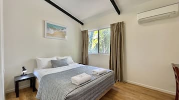 A bedroom with a bed, a nightstand, a painting, a window with curtains, and an air conditioning unit.