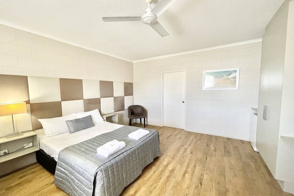 Ein Hotelzimmer mit einem Bett, einem Sessel, einem Schreibtisch, einer Lampe, einem Deckenventilator und einer Tür.