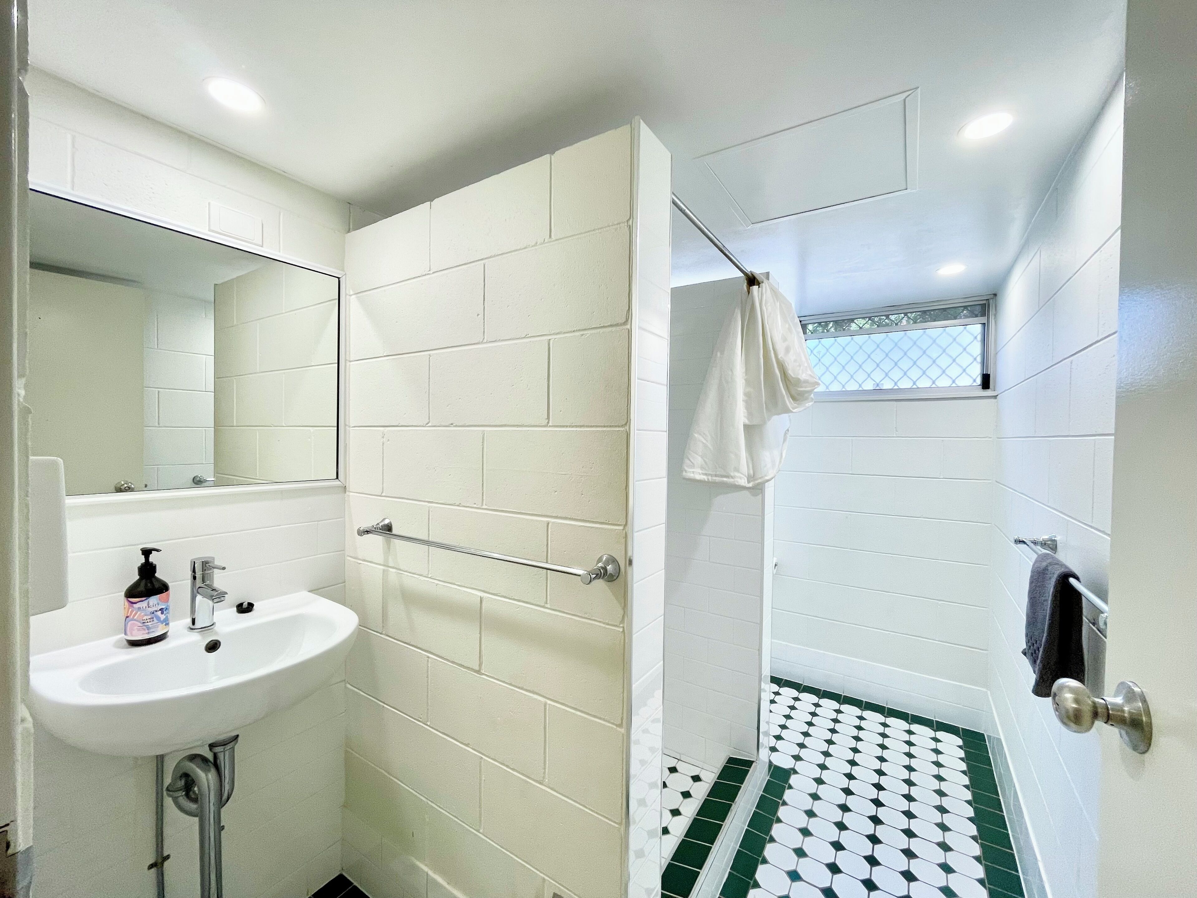 A bathroom with white tiles, a sink with a soap dispenser, a towel rack, and a shower with a patterned floor.