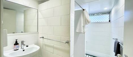 Un baño con azulejos blancos, un lavabo con dispensador de jabón, un perchero para toallas y una ducha con piso estampado.