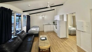 Een knus woongedeelte met een zwart leren bankje, een klein geweven salontafeltje, een kitchenette met witte kastjes en een plafondventilator.