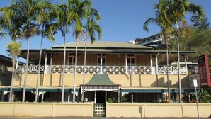 Een hotel in traditionele bouwstijl, met een veranda voorzien van een gedetailleerd hekwerk, diverse palmbomen en een helderblauwe lucht.