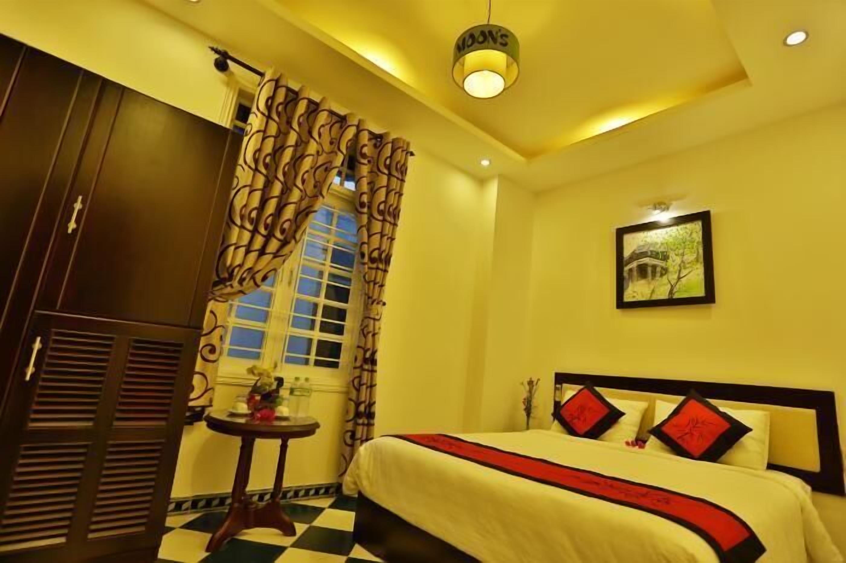 A hotel room with a bed, a small table with a vase, a window with curtains, and a framed picture on the wall.