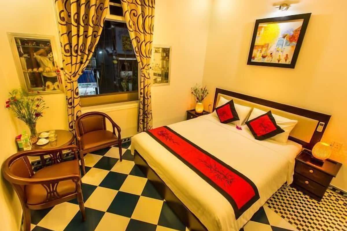 A hotel room with a checkered floor, a bed with red and black bedding, a wooden chair, a small table with a vase, and a window with curtains.