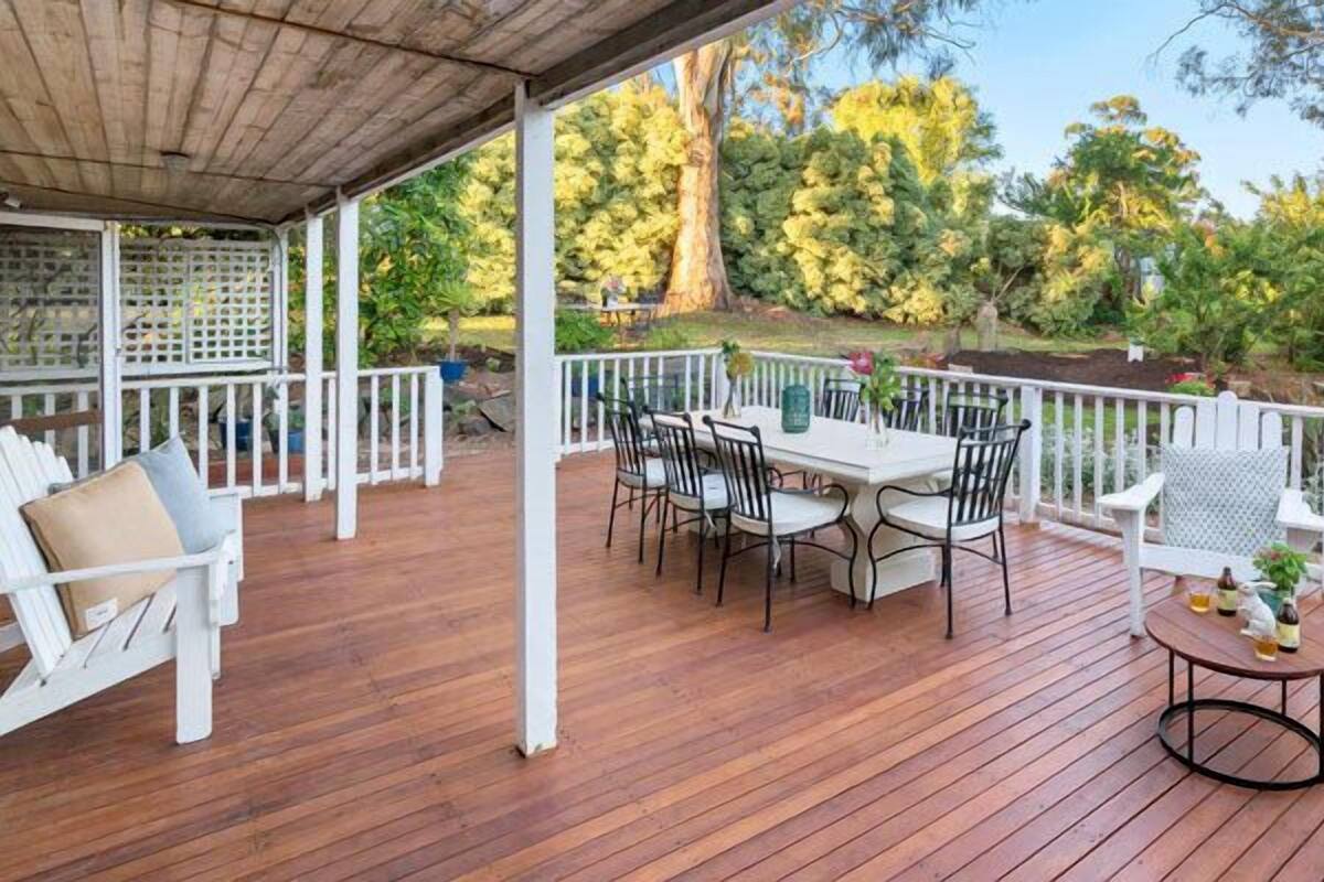 Una terraza exterior techada con piso de madera, muebles blancos y vistas a un jardín con árboles y arbustos.