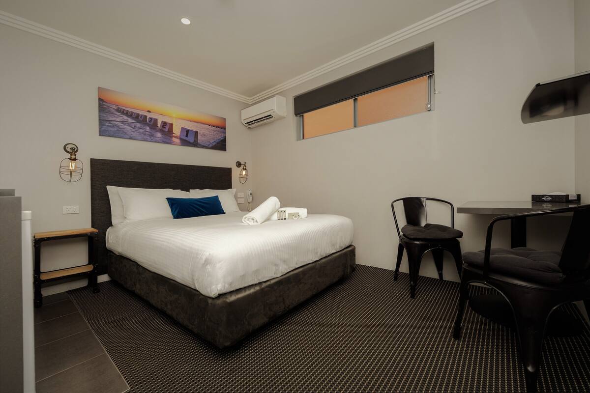 A hotel room with a large bed, a desk, a chair, a wall-mounted air conditioner, and a painting of a sunset over a bridge.