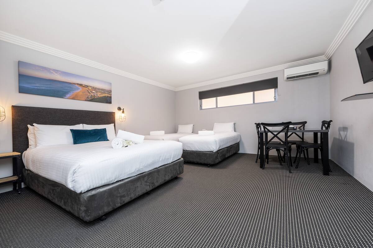 A hotel room with two beds, a desk, and a chair. There is a painting on the wall and an air conditioning unit on the ceiling.