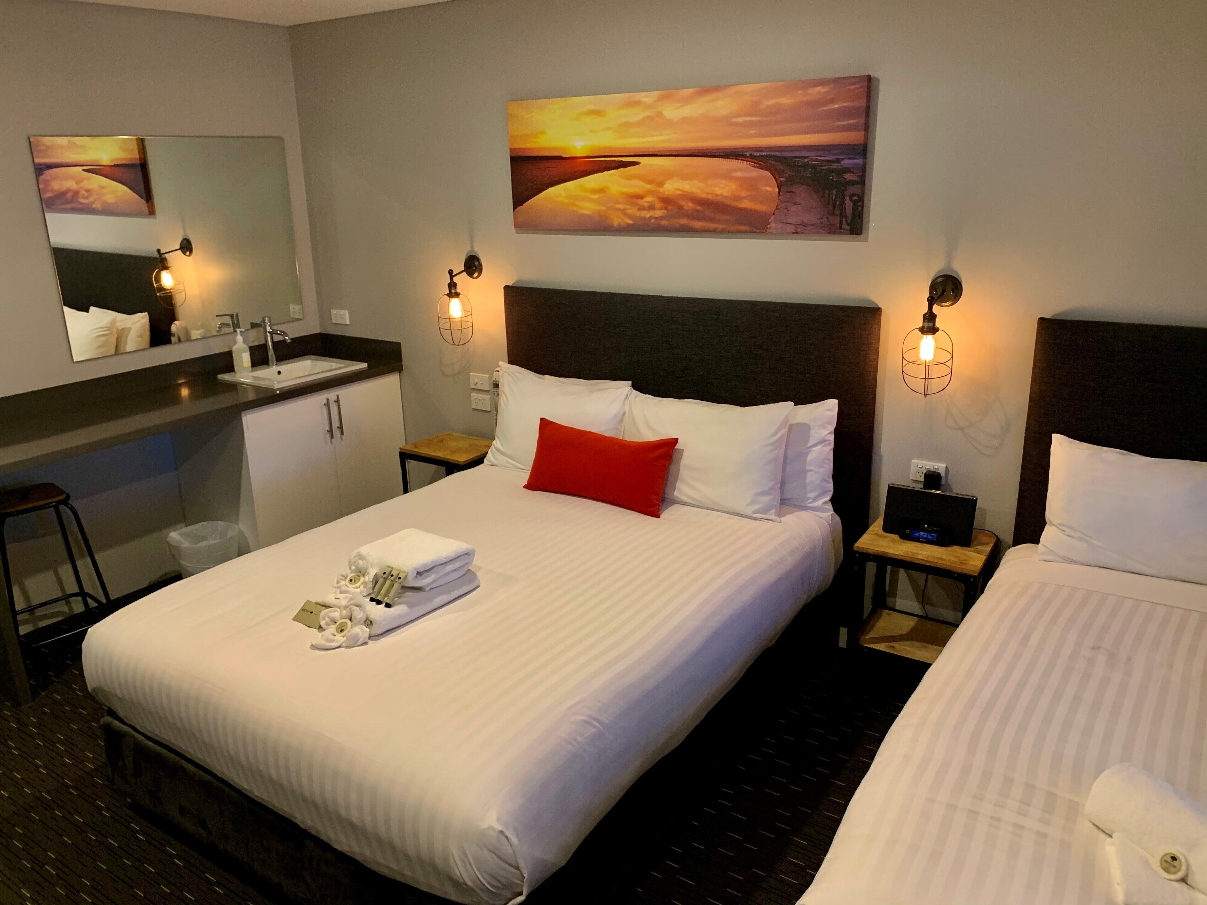 A hotel room with a large bed, a bedside table with a lamp, a small desk with a chair, a mirror, and a painting of a sunset.