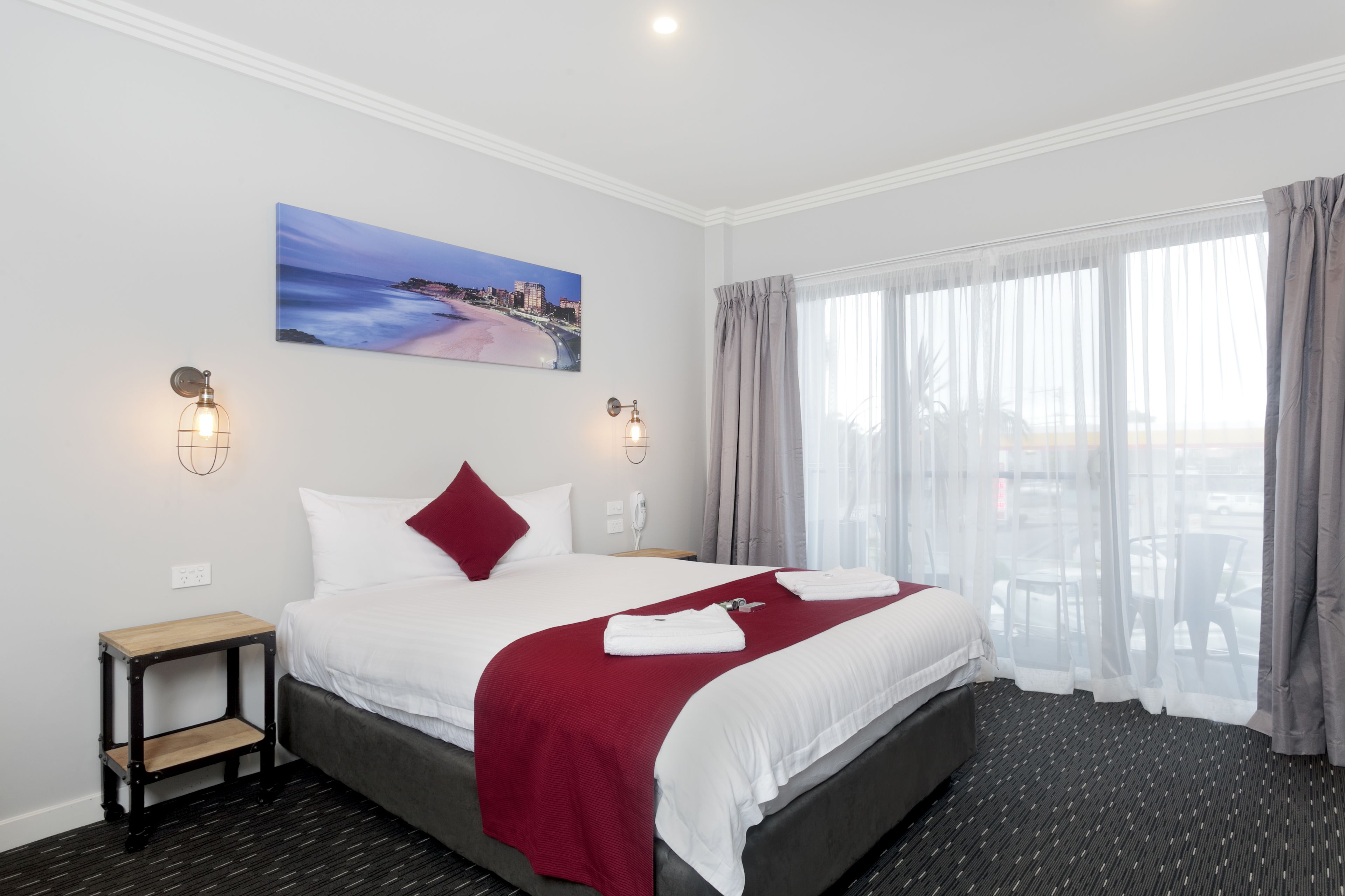 A hotel room with a large bed, a red throw blanket, a painting of a beach, and a view of the outdoors through sheer curtains.