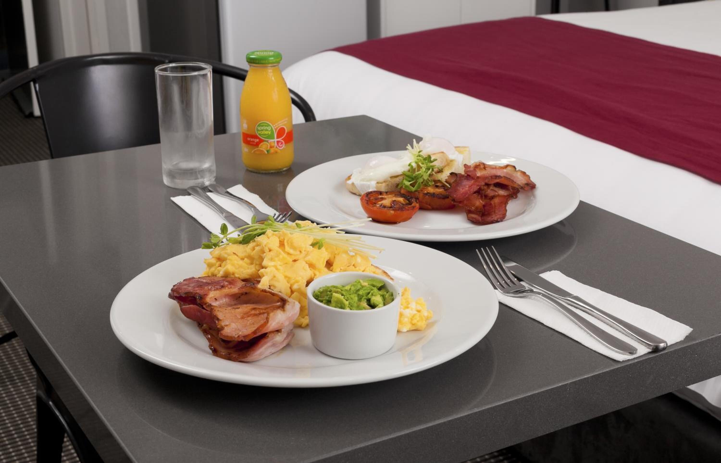 Un desayuno servido con dos platos de comida, incluyendo huevos, tocino, aguacate y huevos revueltos, sobre una mesa con un vaso de agua y una botella de jugo.