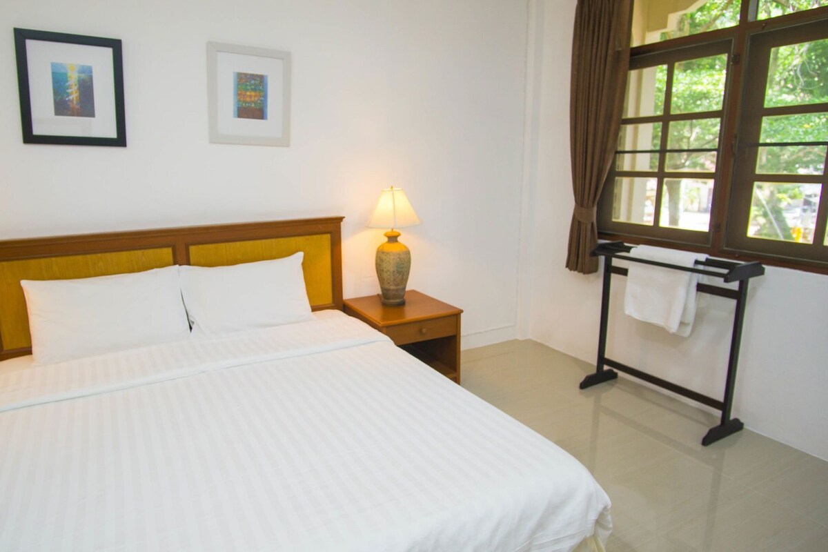A hotel room with a bed, two bedside lamps, a window with curtains, and two framed pictures on the wall.