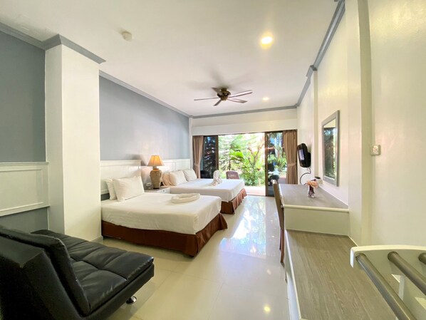 A hotel room with two beds, a ceiling fan, a TV, and a balcony with a view of greenery. - Garden Home Kata (Karon)