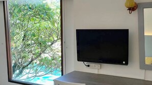Een kamer met een groot raam, een flatscreen televisie aan de muur, een wit bureau en een witte stoel.