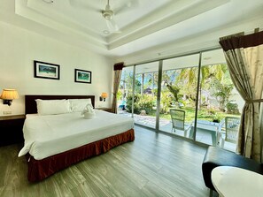 Habitación de hotel con cama, dos lámparas fijadas a la pared, ventilador de techo y un amplio ventanal corredero que da a un espacio exterior con plantas y vistas a una masa de agua.