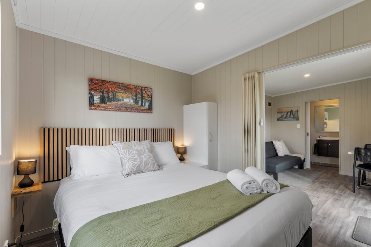 Una habitación de hotel con cama, mesita de noche, lámpara, un cuadro en la pared y un baño visible a través de una puerta abierta.