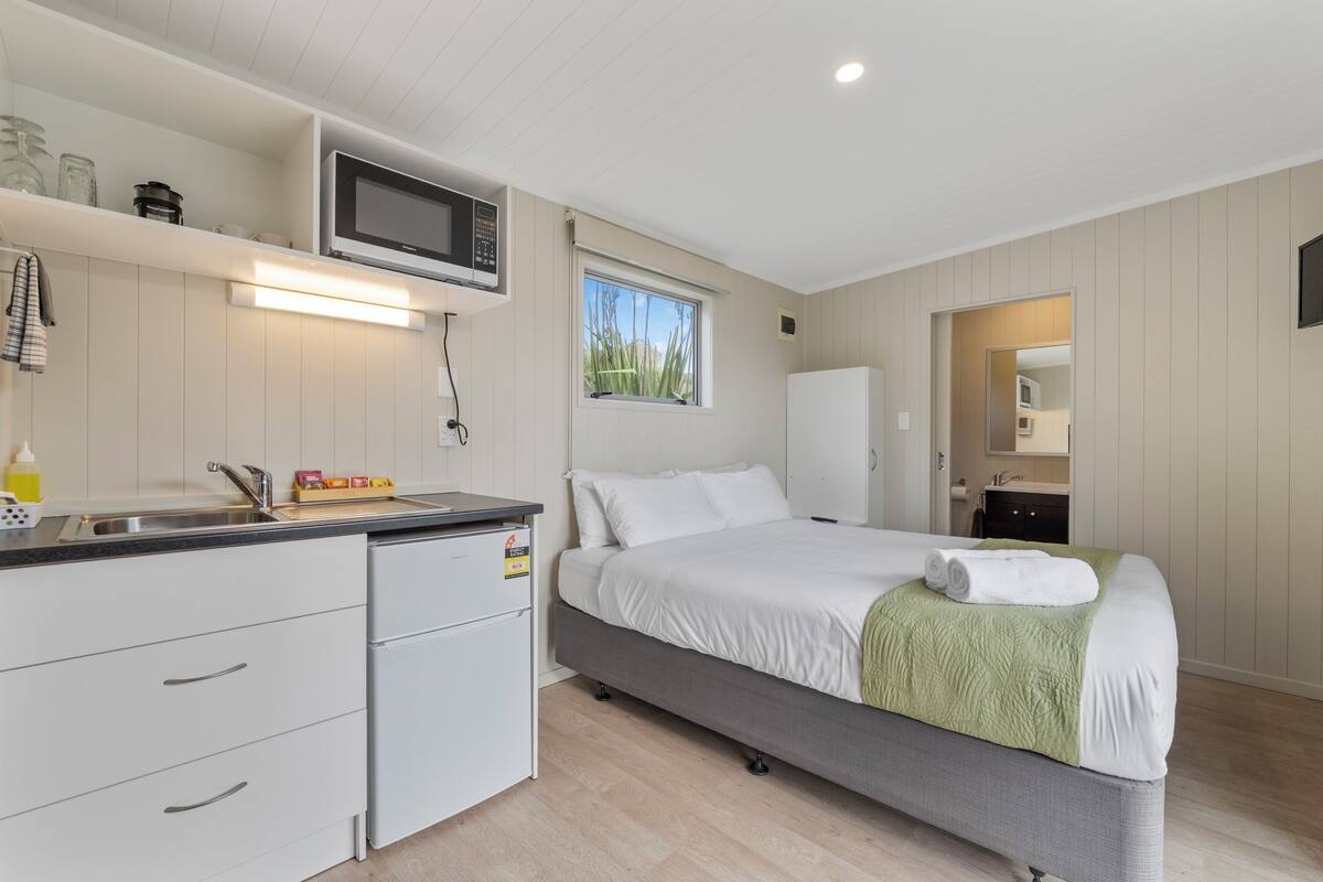 Habitación de hotel compacta con kitchenette, cama con manta verde, ventana con vista y espejo en el baño.