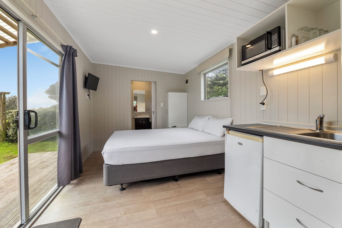 Un dormitorio con cama, una kitchenette pequeña, televisor y puerta corrediza de vidrio a un espacio exterior.