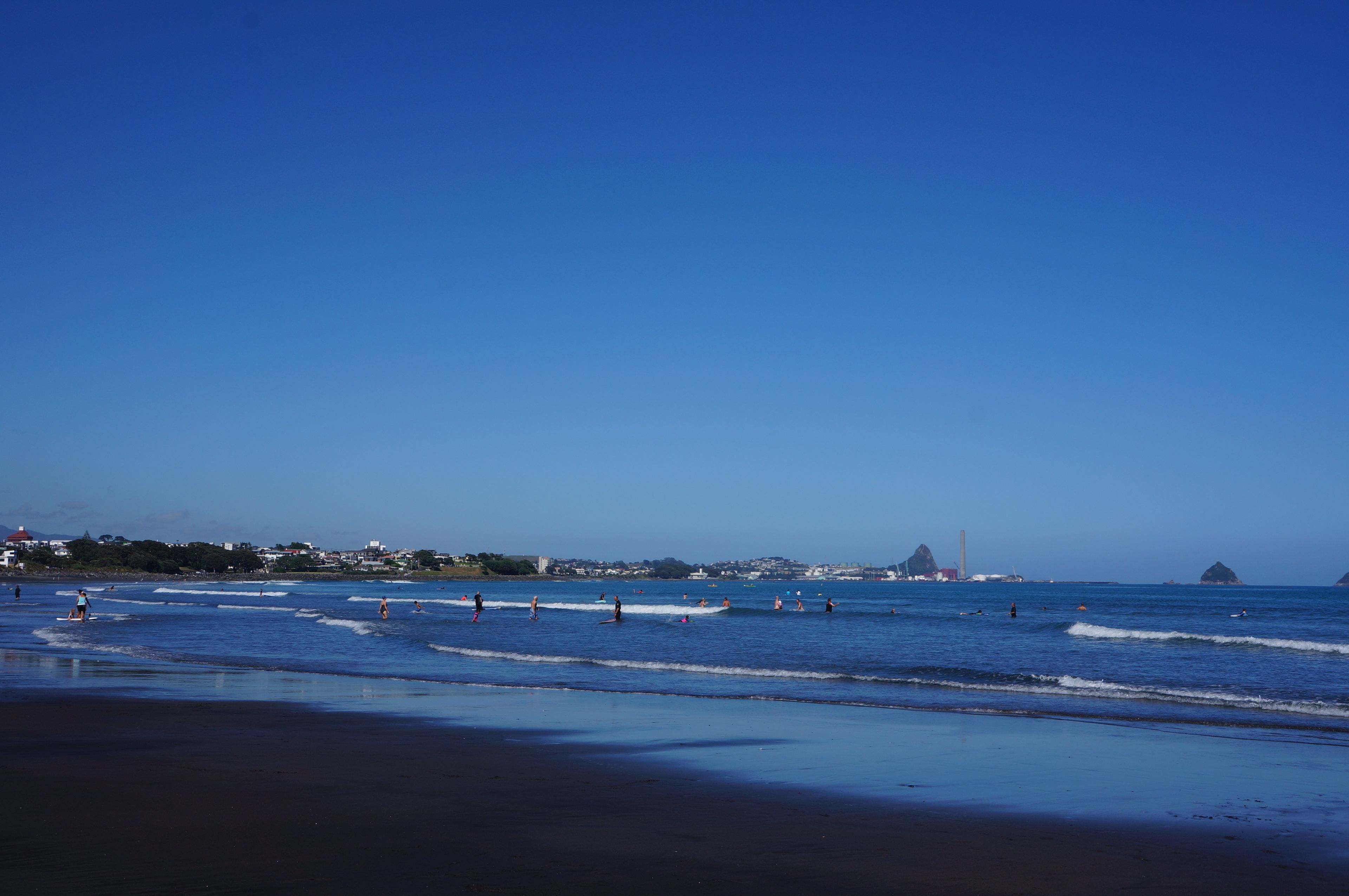 A beach with waves, a clear sky, and distant buildings.