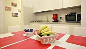 Une cuisine moderne avec des meubles blancs, une machine à café rouge et un four à micro-ondes. On y trouve une table recouverte d’une nappe rouge et blanche, ainsi qu’un panier rempli de bananes et de pommes.