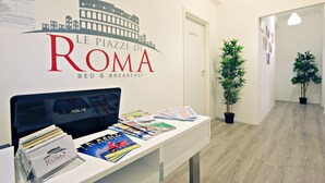 Un comptoir d’accueil avec une pancarte indiquant « Le Piazza Di Roma Bed & Breakfast », un écran d’ordinateur, des brochures et une plante en pot.