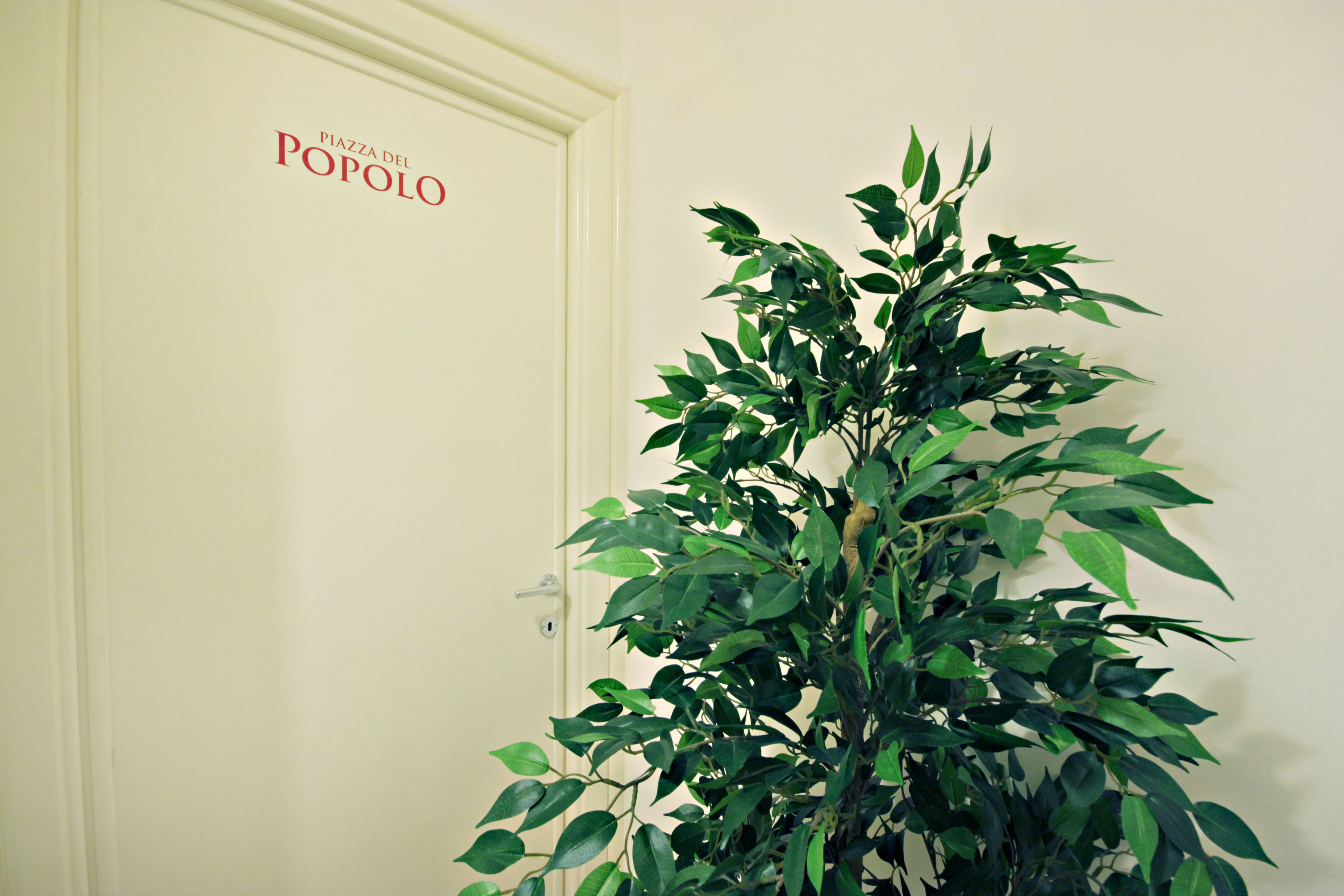 Une porte sur laquelle est apposée une plaque indiquant « Piazza del Popolo », et une grande plante en pot placée à côté.