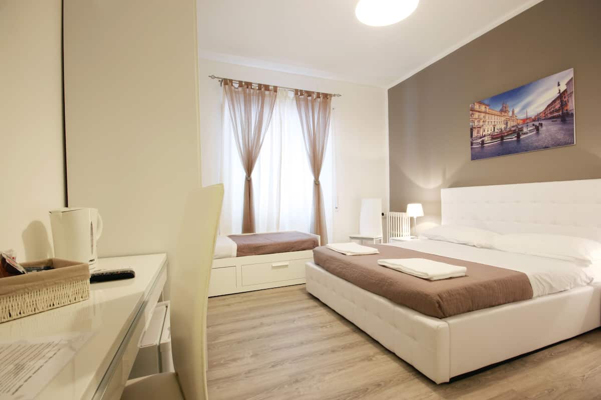 A hotel room with a bed, a desk with a chair, a lamp, and a window with curtains.