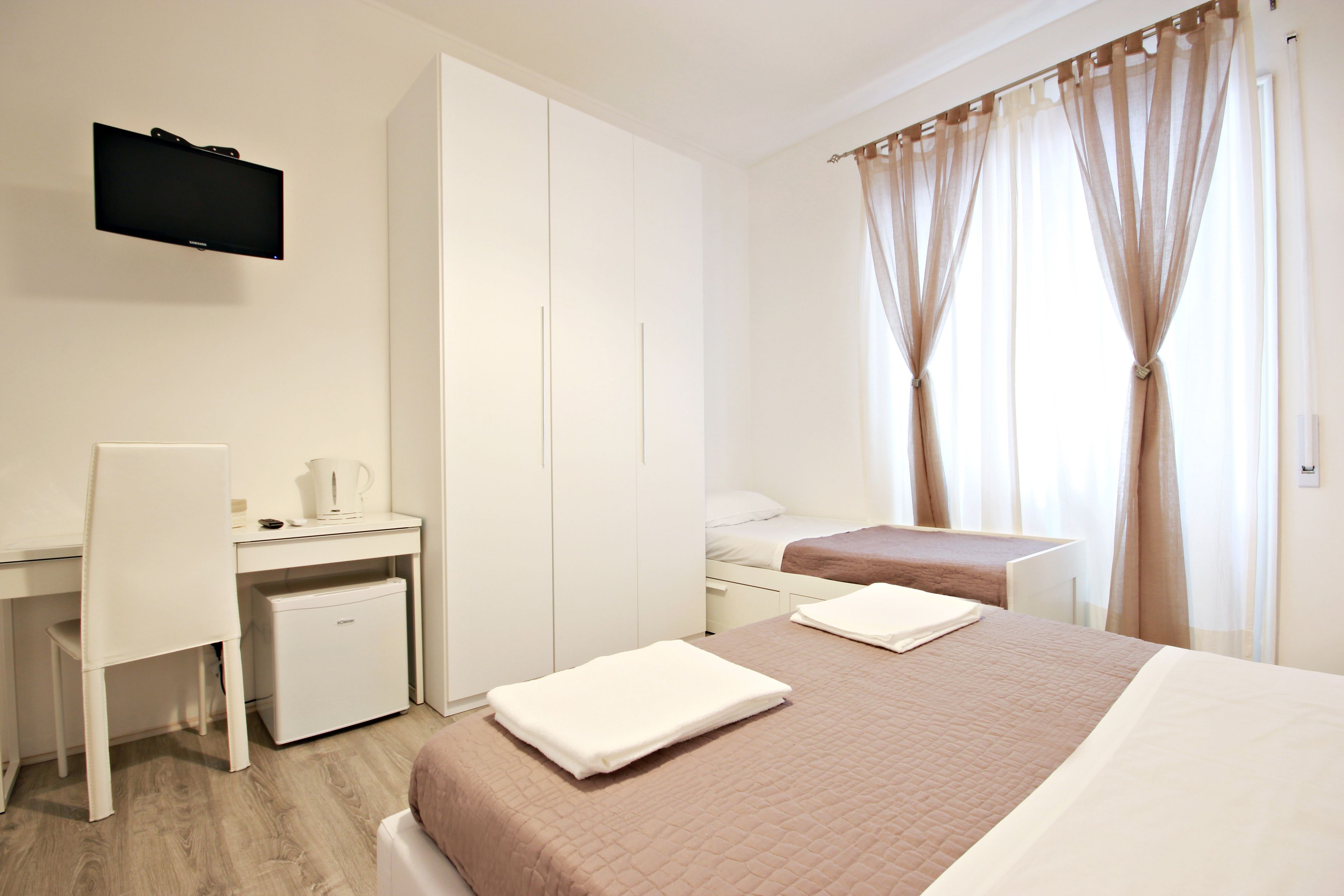 Habitación de hotel con cama, escritorio, silla, refrigerador pequeño y televisor montado en la pared.