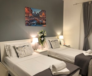 A hotel room with two beds, a nightstand with a lamp, a vase with flowers, and a painting of a cityscape on the wall. - Le Piazze di Roma B&B (Rome)