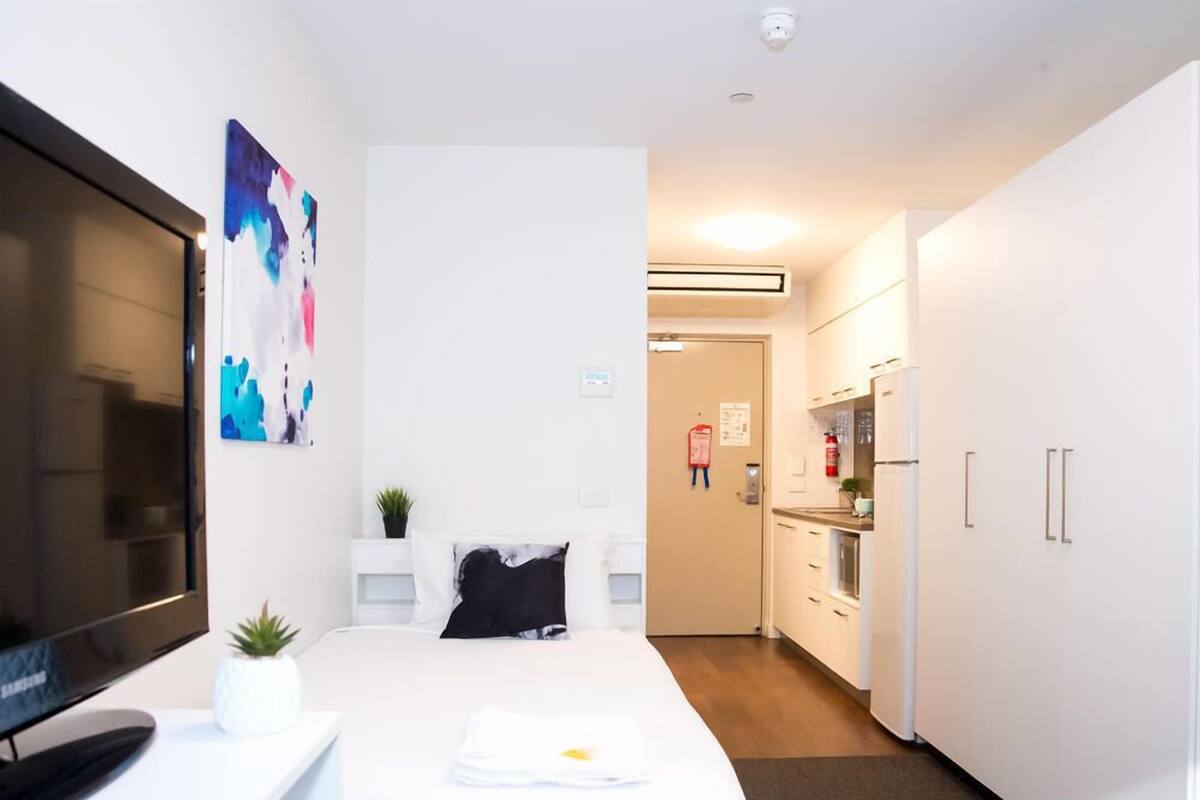 A hotel room with a bed, a TV, a painting, a small plant, and a kitchenette.