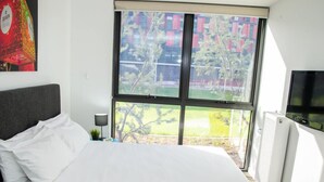 A modern hotel room with a large bed, a small table with a lamp, a TV, and a window with a view of buildings and trees. - Deakin Residential Services (Burwood)