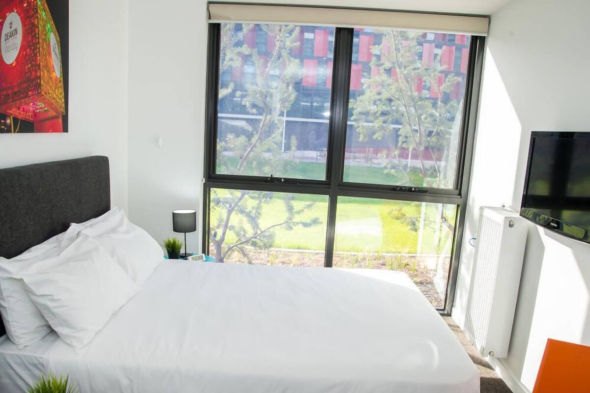 A modern hotel room with a large bed, a small table with a lamp, a TV, and a window with a view of buildings and trees.