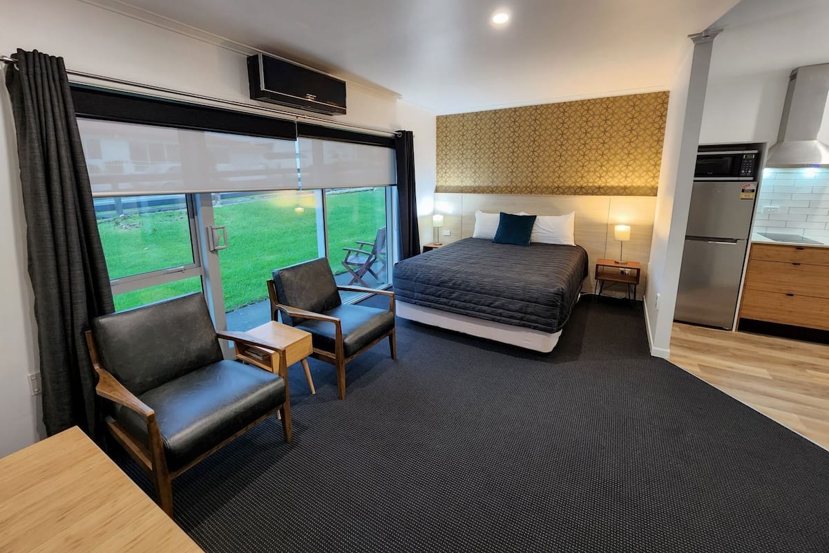 A modern hotel room with a bed, a desk, two chairs, a kitchen area, and a large window with a view.
