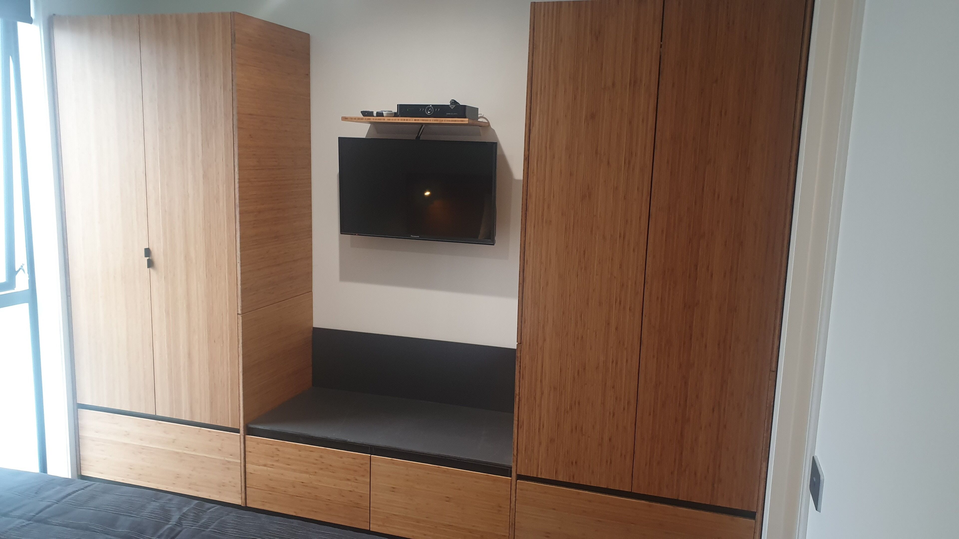 A room with a built-in TV on a wooden wall, a small shelf above it, and a bench with drawers.