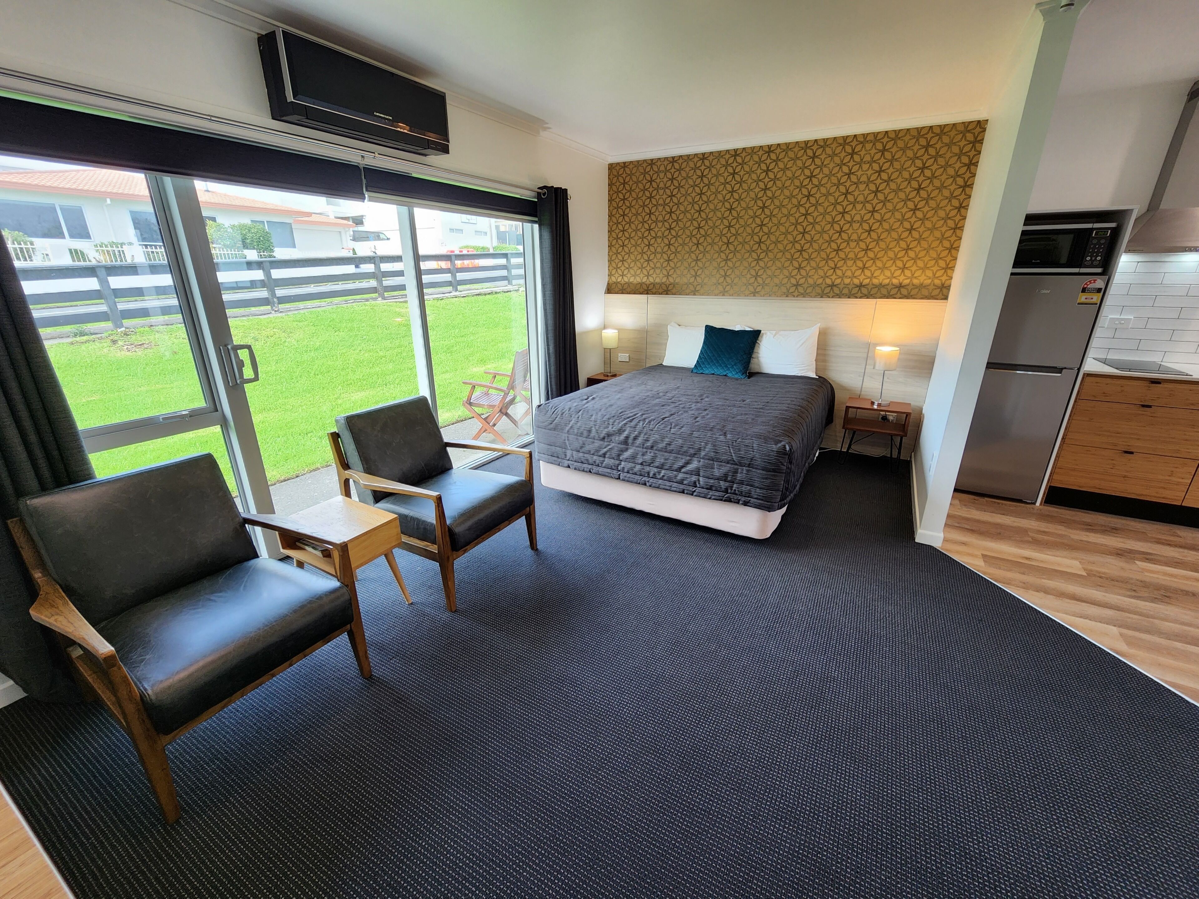 Una habitación de hotel moderna con una cama grande, una zona de estar con una silla y una mesita, una kitchenette y vistas a un césped verde y edificios.