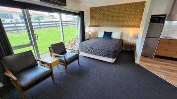 Una habitación de hotel moderna con una cama grande, una zona de estar con una silla y una mesita, una kitchenette y vistas a un césped verde y edificios.