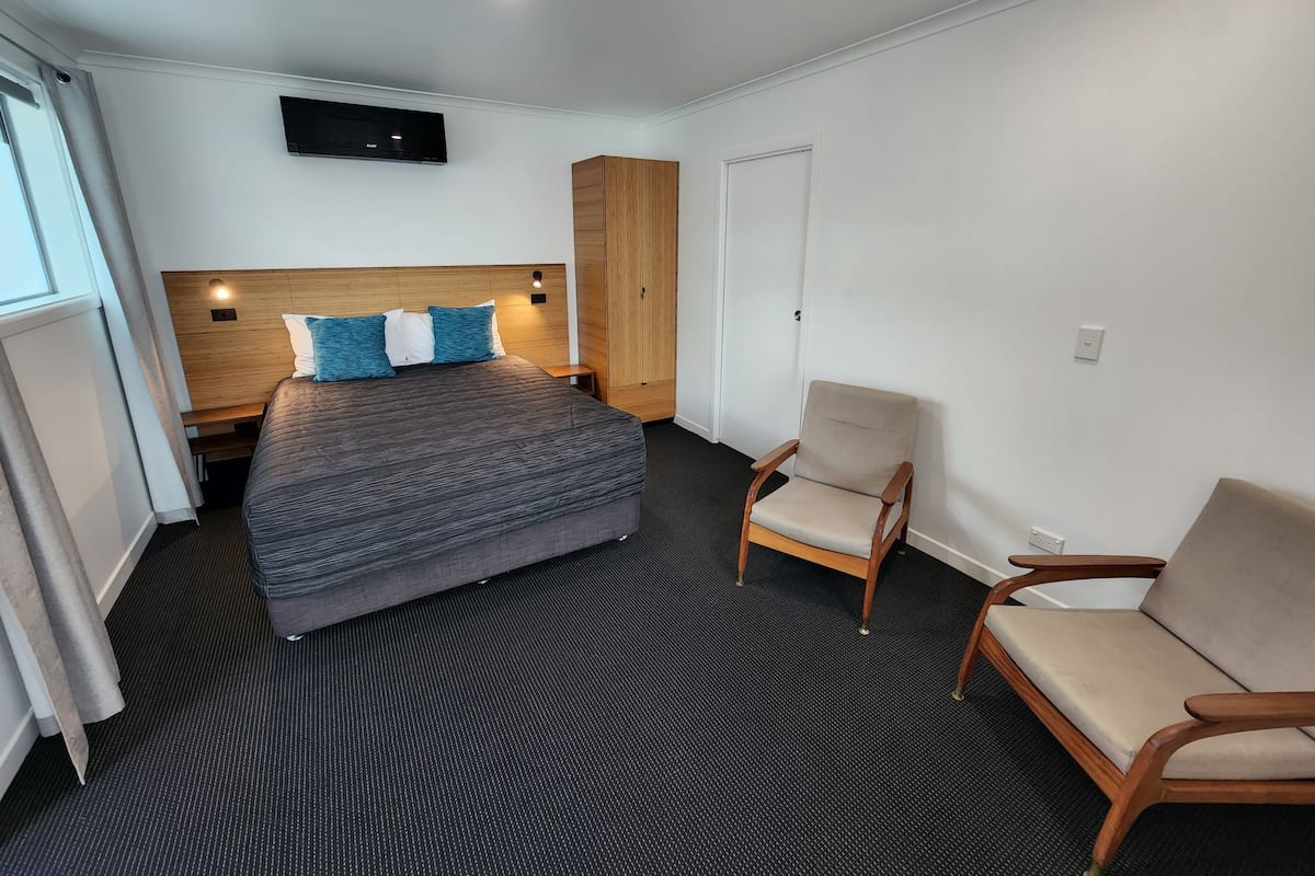 A hotel room with a bed, a wooden wardrobe, a chair, and a television mounted on the wall.