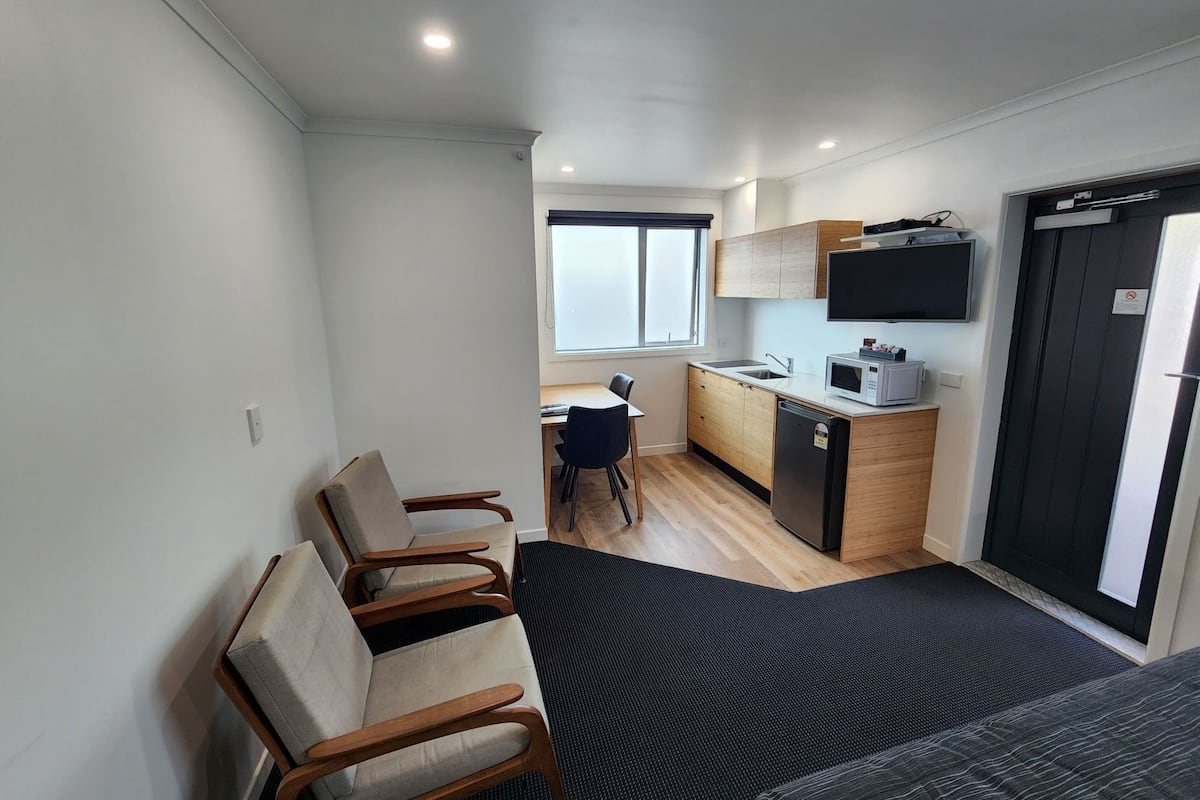A compact room with a kitchenette, a dining area with chairs, a bed, and a large window.