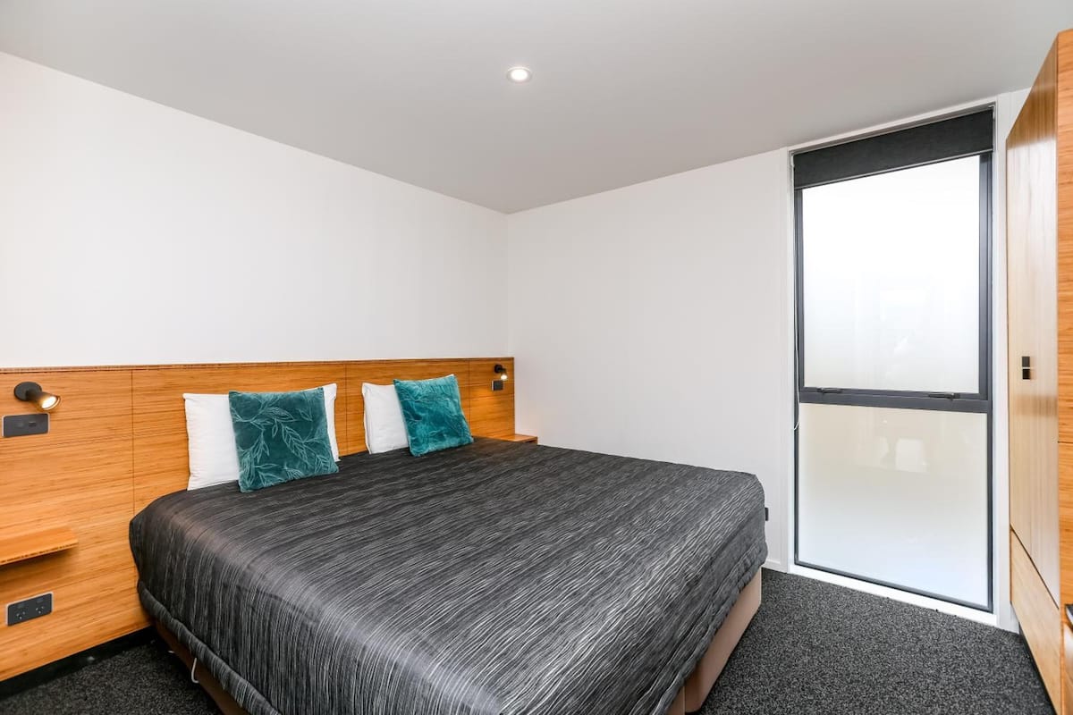A modern hotel room with a large bed, a wooden headboard, and a sliding glass door to the outside.