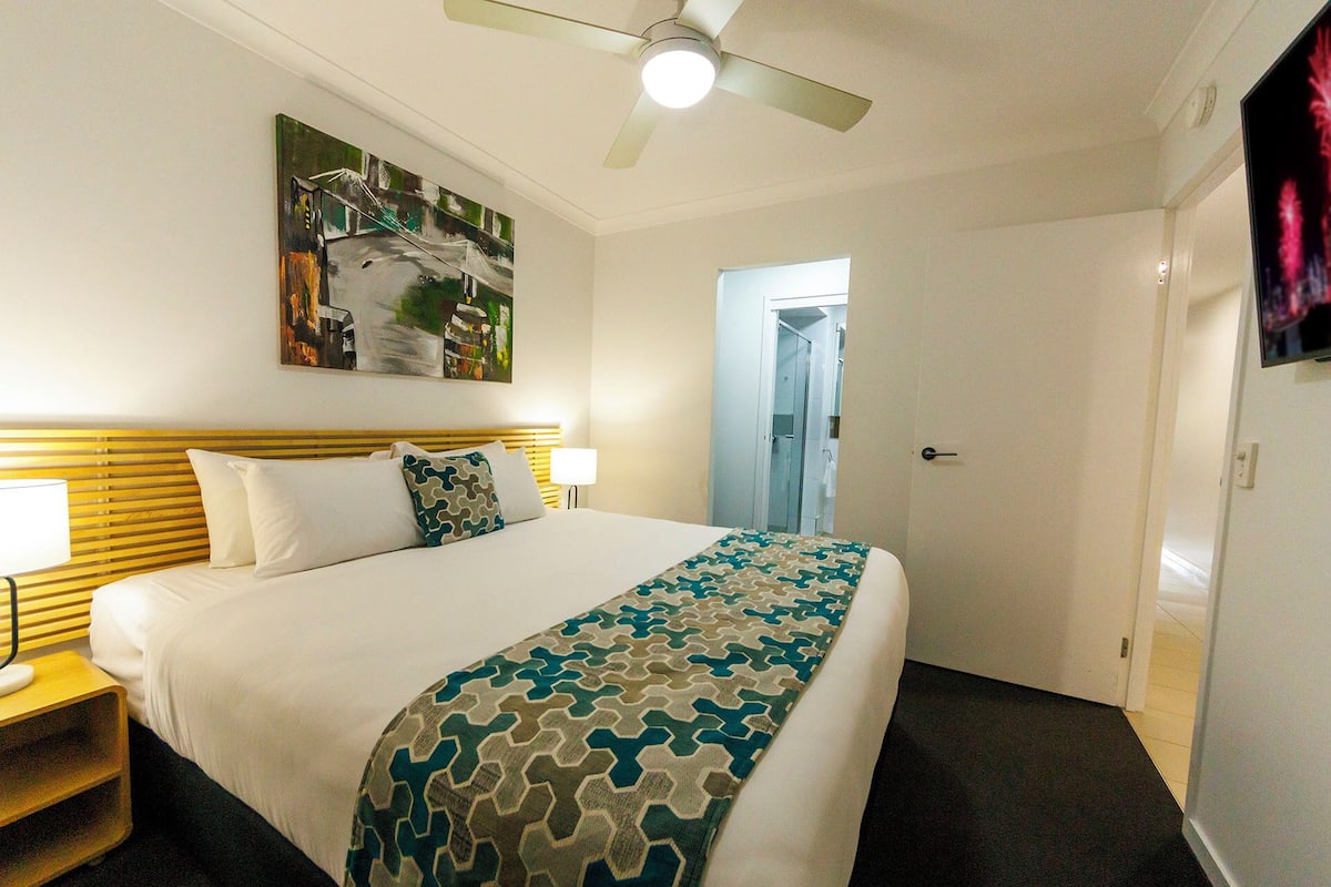 Ein Hotelzimmer mit einem Bett, einem Deckenventilator, einem Gemälde an der Wand und einer Tür zu einem weiteren Raum.