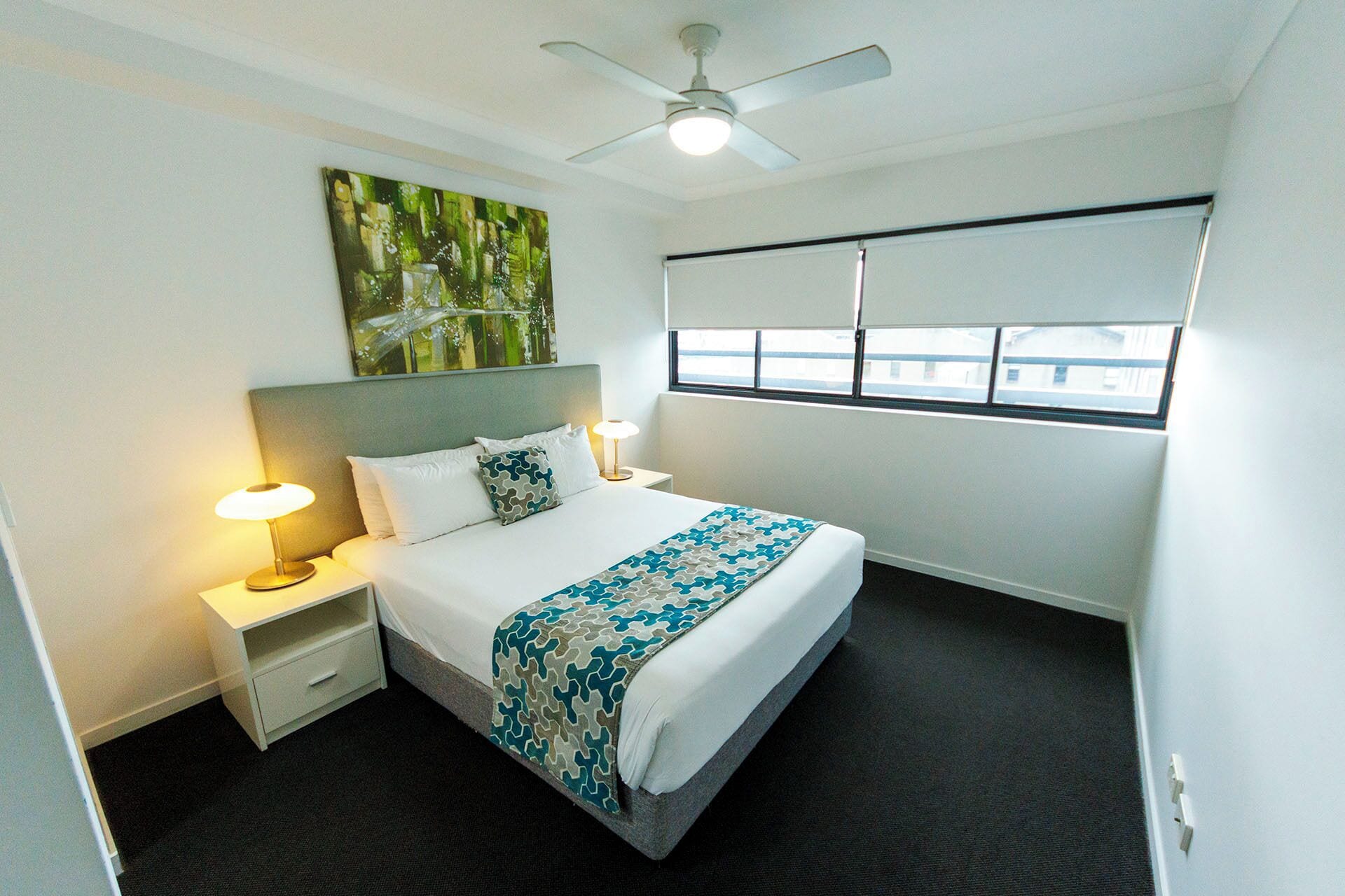 A hotel room with a bed, two bedside lamps, a ceiling fan, a window with blinds, and a painting on the wall.