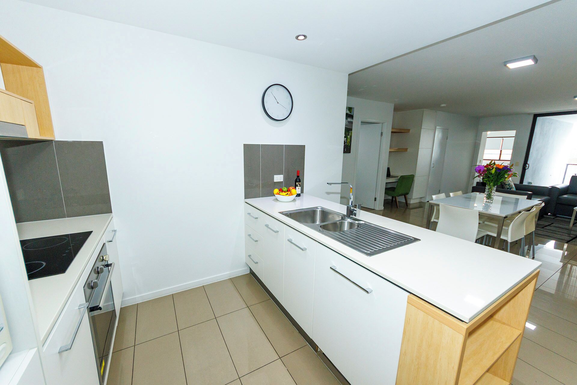 A modern kitchen with white cabinetry, a built-in oven, a stainless steel sink, and a clock on the wall.
