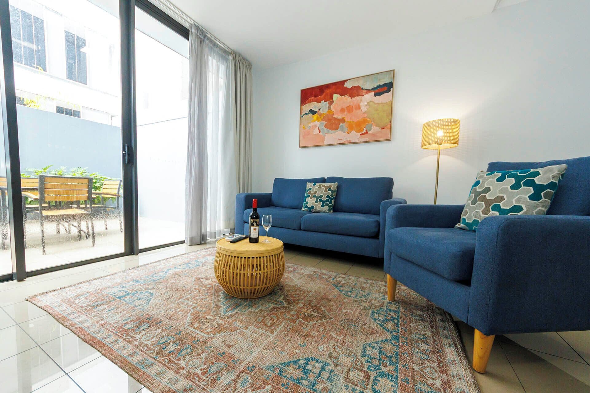 A living room with a blue sofa, a round wooden coffee table, a colorful rug, a floor lamp, and a painting on the wall.