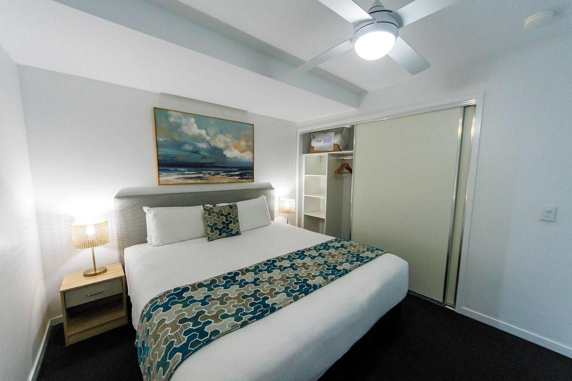 Une chambre d’hôtel avec un lit, un ventilateur de plafond, un tableau, une armoire et une table de chevet avec une lampe.
