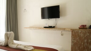 A hotel room with a bed, a TV mounted on a wooden shelf, a small table with a kettle and cups, and a window with curtains. - Sugiras Living (Denpasar)