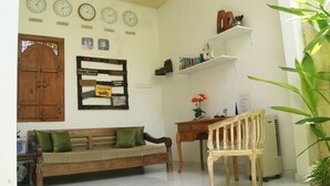 A living room with a wooden sofa, a wooden chair, a desk with a computer, shelves with books and decorative items, and a wall clock with multiple time zones. - Sugiras Living (Denpasar)