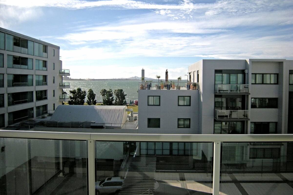 Un edificio moderno con balcón de cristal con vistas a una masa de agua y un horizonte urbano distante.